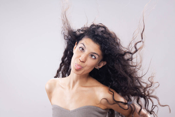 Beautiful brunette girl with long curly hair in the air, studio portrait. Funny clumsy face expression. - Foto, Imagen