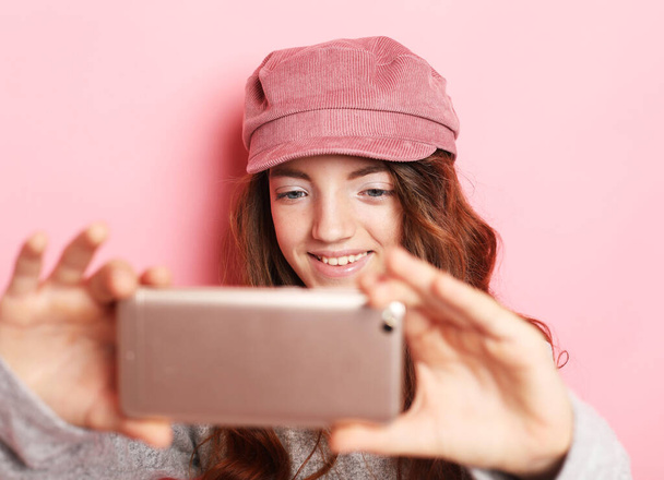 Lifestyle, tehnology and people concept: Portrait of a cheerful little girl wearing hat taking a selfie isolated over pink background - Foto, immagini