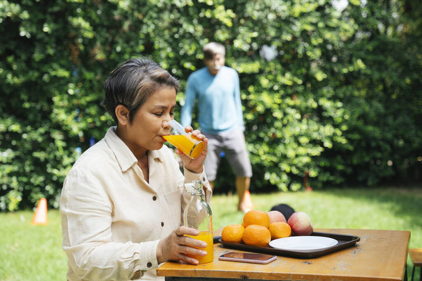 Ηλικιωμένη γυναίκα απολαμβάνουν πόσιμο χυμό πορτοκαλιού με γυαλί σε εξωτερικούς χώρους στο πάρκο. - Φωτογραφία, εικόνα