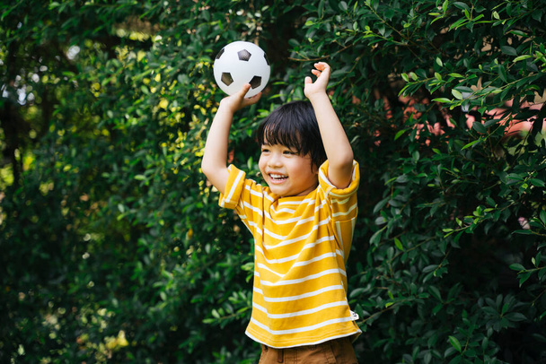 Wesoły azjatycki chłopiec trzyma piłkę nożną pokazując ręce w powietrzu w parku. - Zdjęcie, obraz