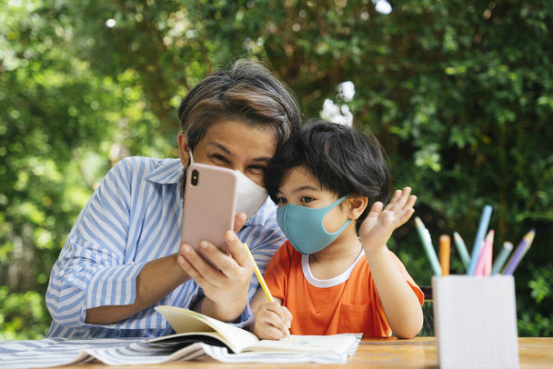 Seniorin trägt Maske mit Smartphone zusammen mit ihrem Enkel. - Foto, Bild