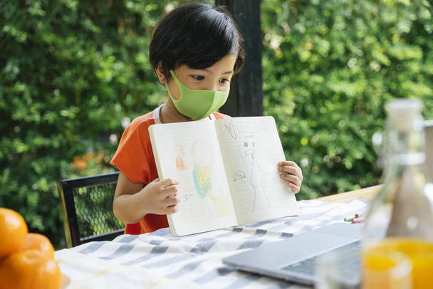 Asyalı çocuk dizüstü bilgisayarda öğretmenine çizim yaparken maske takıyor. Evden ders çalış.. - Fotoğraf, Görsel
