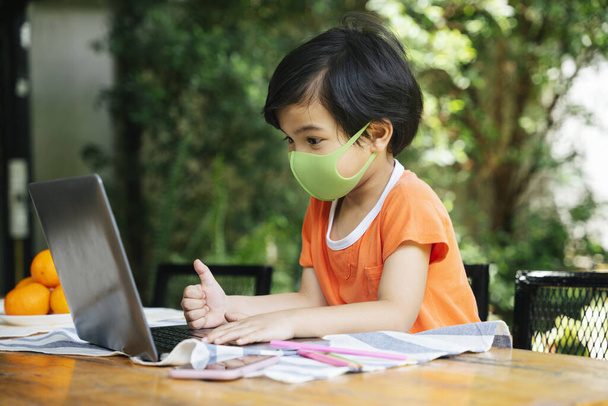 Vista laterale - ragazzo asiatico indossare maschera utilizzando computer portatile in cortile. Studiare da casa. - Foto, immagini