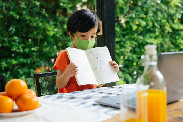 Azjatyckie dziecko nosi maskę, rysując dzieła sztuki w książce dla nauczyciela na laptopie. Badanie z domu. - Zdjęcie, obraz