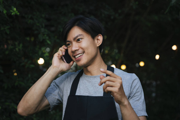 alegre asiático camarero llevar negro delantal hablando en el teléfono en patio al aire libre. - Foto, Imagen