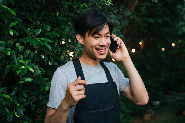 Veselý asijský číšník nosit černou zástěru mluvit do telefonu na dvoře venku. - Fotografie, Obrázek