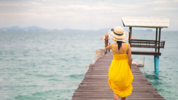 De cerca retrato de nuevo joven mujer asiática en un vestido amarillo tocando un sombrero en su cabeza caminando sobre un puente de madera en la playa con un fondo de cielo, espacio de copia fpr lado izquierdo en blanco - Foto, Imagen