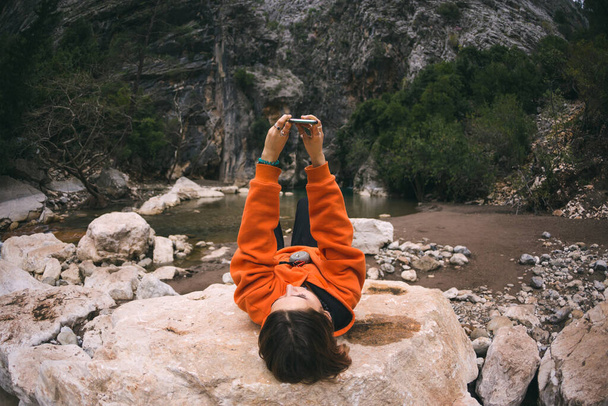 Μια γυναίκα βρίσκεται σε μια μεγάλη πέτρα και παίρνει μια selfie σε ένα smartphone, ένα κορίτσι φωτογραφίζεται σε ένα φόντο των όμορφων βραχώδη βουνά, Ταξιδεύοντας σε γραφικά σημεία. - Φωτογραφία, εικόνα