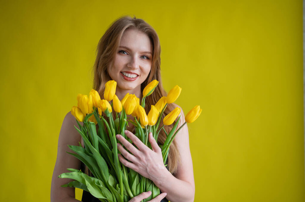 Caucasian woman with an armful of yellow tulips on a yellow background. International Womens Day. Bouquet of spring flowers - Foto, Imagen