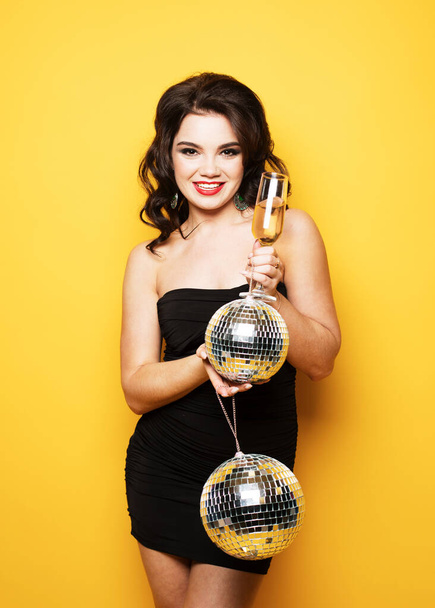 Christmas, celebration party, holiday concept. Young brunette woman in evening dress holding champagne and disco ball. Brighrt make up and Wavy hairstyle. Over yellow background. - Foto, immagini