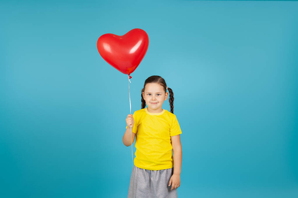 cute, demure girl with pigtails in a yellow T-shirt holds a flying red balloon in the shape of a heart , isolated on a blue background - Foto, Imagem