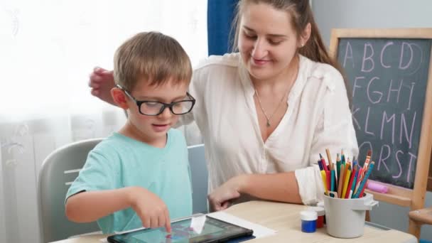Gözlüklü çocuk tablet bilgisayarda oynuyor. Anne küçük oğluna sarılıp ödev yaparken gadet kullanıyor. Çocukların görme ve görme sorunları var. - Video, Çekim