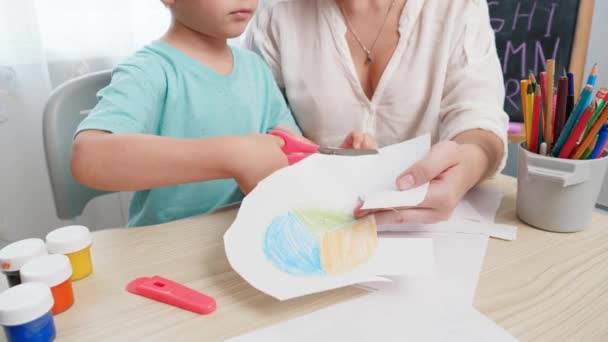 Kleine jongen knipt getekend beeld uit op papier met een schaar. Moeder die haar zoontje helpt. Kleuteronderwijs en -leren thuis - Video