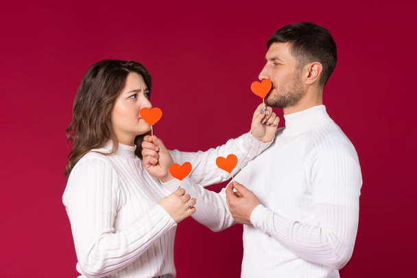 Photo of young couple joking, red background on St.Valentine's Day. Closes their mouths with handmade paper hearts on toothpick. High quality photo - Valokuva, kuva
