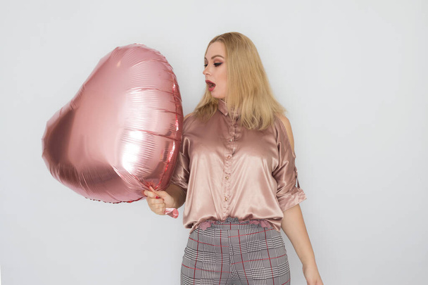 Blonde business woman in pink blouse holding big pink heart balloon in her hands over white background in studio - Valokuva, kuva