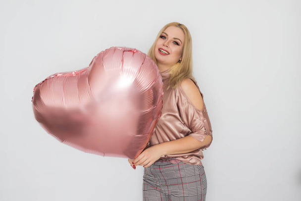 Blonde business woman in pink blouse holding big pink heart balloon in her hands over white background in studio - Foto, Imagen