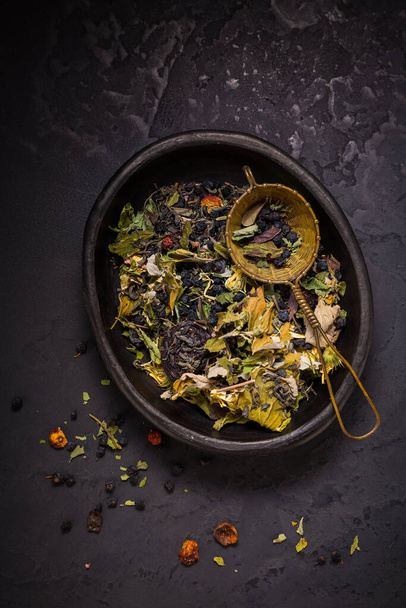 Mixture of teas, dried organic tea herbs and fruits in bowl with tea strainer on black background - Photo, Image
