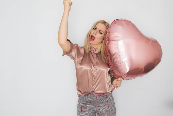 Blonde business woman in pink blouse holding big pink heart balloon in her hands over white background in studio - Zdjęcie, obraz