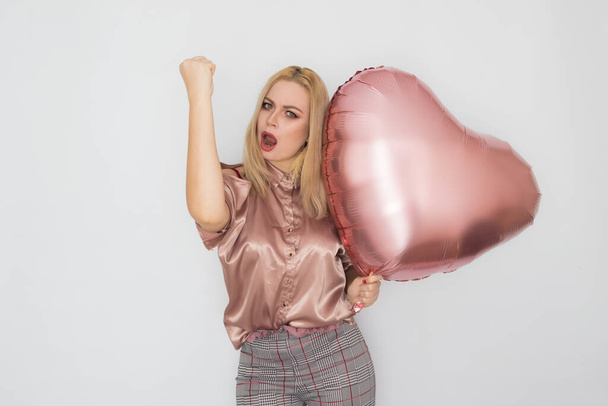 Blonde business woman in pink blouse holding big pink heart balloon in her hands over white background in studio - Фото, изображение
