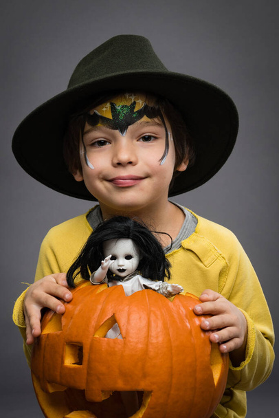 Menino alegre um chapéu mantém uma abóbora com a boneca assustadora em um fundo cinza. Halloweencelebration. - Foto, Imagem