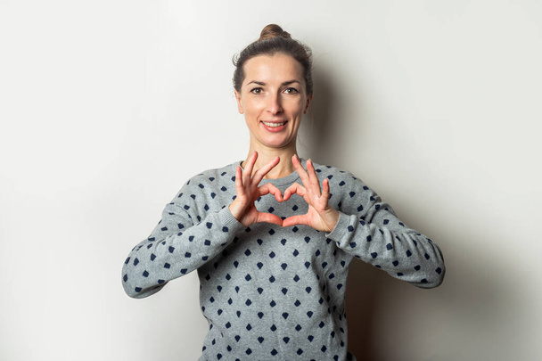 Young woman in a sweater makes a heart gesture on a light background. Valentine's Day concept. Banner. - Photo, Image