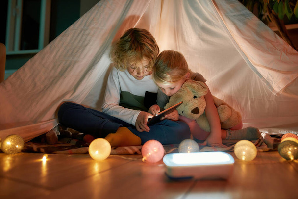Dos hermanitos, hermano y hermana mirando enfocados mientras usan tableta digital, sentados en una cabaña hecha con sábanas - Foto, imagen