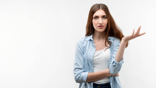 Displeased girl with long chestnut hair, dressed in casual clothes, is shrugging shoulders being confused, gesturing with raised palm, confused with question. Human emotions concept - Foto, Bild