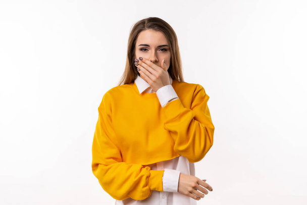Frustrated brunette girl in casual clothes is covering mouth with hand not willing to disclose something important. Keep a secret. Studio shot, white background - Photo, Image