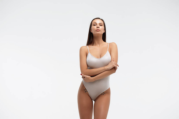 Tender vrouwelijk dragen bodysuit staande met gevouwen armen op witte achtergrond in studio en wegkijkend - Foto, afbeelding