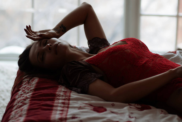 Vue latérale de la jeune femme sexuelle avec de longs cheveux foncés dans un soutien-gorge rouge élégant couché sur un lit confortable avec les yeux fermés près de l'arbre de Noël décoré - Photo, image