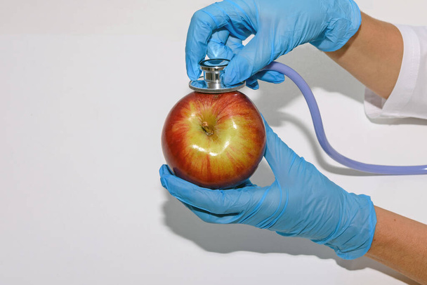 Handen in medische handschoenen houden een appel vast, die wordt onderzocht met een stethoscoop. Concept van het probleem van de ecologie en het gebruik van meststoffen. Sluiten.. - Foto, afbeelding