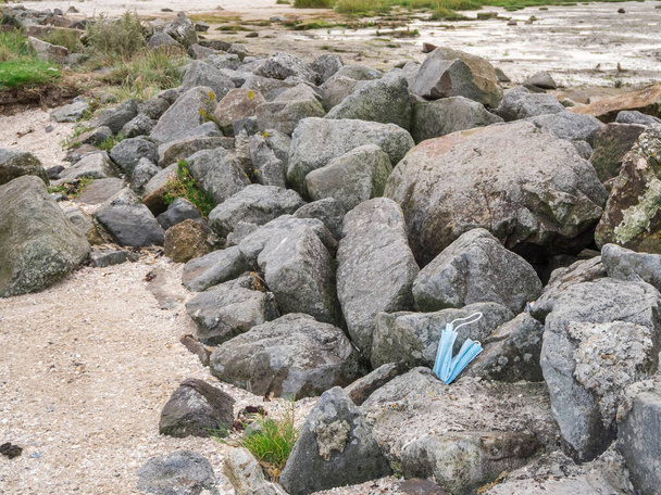 A light blue mouth and nose mask lying on boulders on the North Sea shore. - Photo, Image