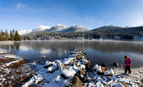 Потрясающая зимняя пейзажная панорама. Замерзшее озеро, снег и покрытые снегом горы. Маленькая девочка в розовом на переднем плане - Фото, изображение
