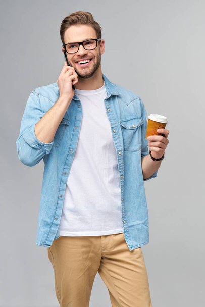 pretty casual man in blue jeans shirt holding his phone and cup of coffee to go standing over studio grey background - Photo, image