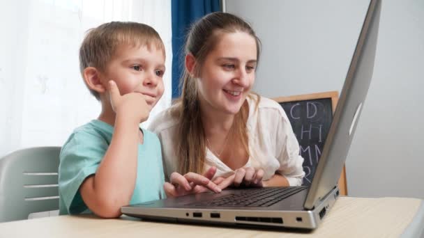 Feliz chico inteligente apuntando a la computadora portátil mientras la madre la usa y teclea en el teclado. Educación infantil y estudio en computadoras en casa - Metraje, vídeo