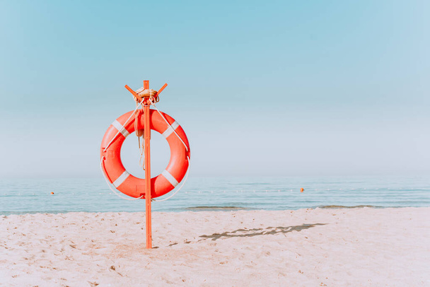 red lifebuoy on a sandy beach in pastel colors. Red lifebuoy on the sandy shore of the beach on a sunny day against the background of blue water - Фото, зображення