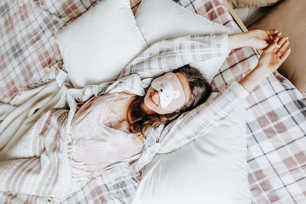 A sleepy brunette girl lies in bed with a blindfold waking up in the morning. Cozy home morning in cute pajamas. Have a good time, relax and dream. Healthy sleep. - Фото, изображение
