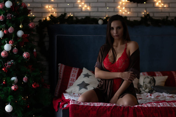 Senhora jovem auto-assegurada em robe de seda marrom e sutiã branco sentado em cama confortável em quarto acolhedor decorado com guirlandas brilhantes e grinalda durante as férias de Natal - Foto, Imagem