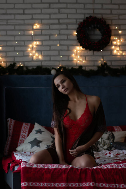 自己保証若いです女性で茶色の絹のローブと白いブラジャーで快適なベッドの上に座っている居心地の良い部屋で輝くガーランドとクリスマス休暇中の花輪で飾られた - 写真・画像