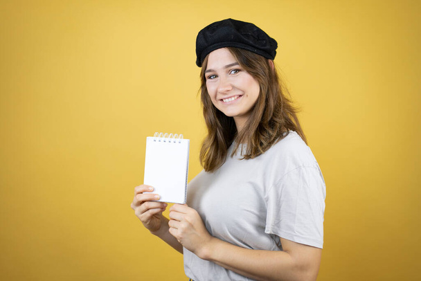Hermosa chica caucásica joven con aspecto francés con boina sobre fondo amarillo aislado sonriendo y mostrando cuaderno en blanco en su mano - Foto, imagen