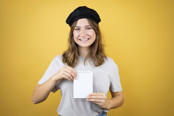 Bela jovem caucasiana vestindo olhar francês com boina sobre fundo amarelo isolado sorrindo e mostrando caderno em branco em sua mão - Foto, Imagem