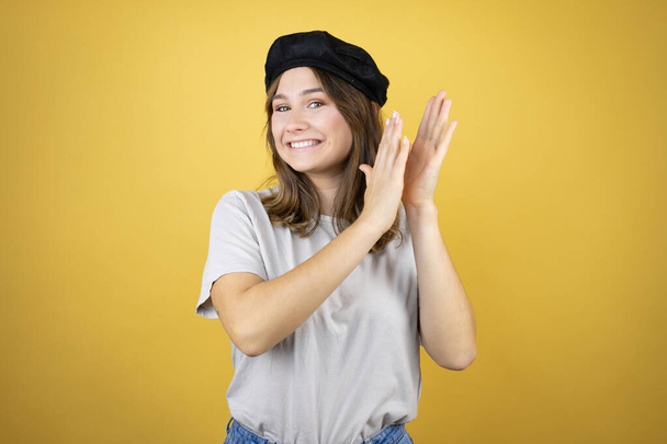 Gyönyörű fiatal kaukázusi lány visel francia megjelenés svájcisapkával felett elszigetelt sárga háttér tapsol és tapsol boldog és örömteli, mosolygós büszke kezét együtt - Fotó, kép