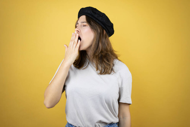 Beautiful young caucasian girl wearing french look with beret over isolated yellow background bored yawning tired covering mouth with hand. Restless and sleepiness. - Valokuva, kuva