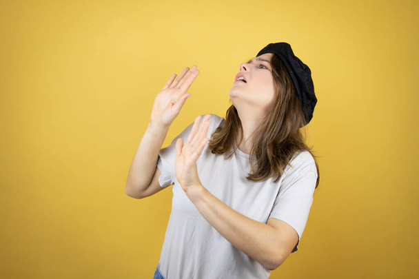 Beautiful young caucasian girl wearing french look with beret over isolated yellow background scared with her arms up like something falling from above - Foto, Imagem