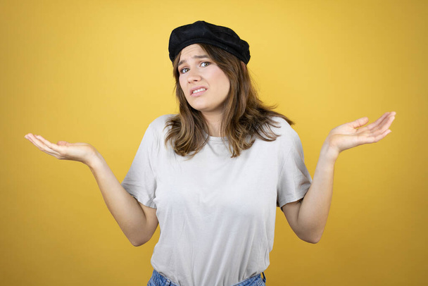 Beautiful young caucasian girl wearing french look with beret over isolated yellow background clueless and confused expression with arms and hands raised - Photo, Image