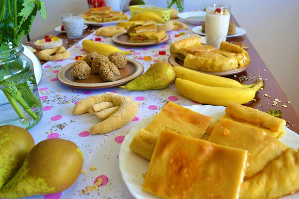 Brunch τραπέζι με λευκό τραπεζομάντηλο με ροζ πουά και πάνω του λουλούδια, πίτα, φρούτα, τσάι από βότανα, σοκολάτα, κεφτεδάκια, κέικ ρυζιού και άλλα νόστιμα πιάτα - Φωτογραφία, εικόνα