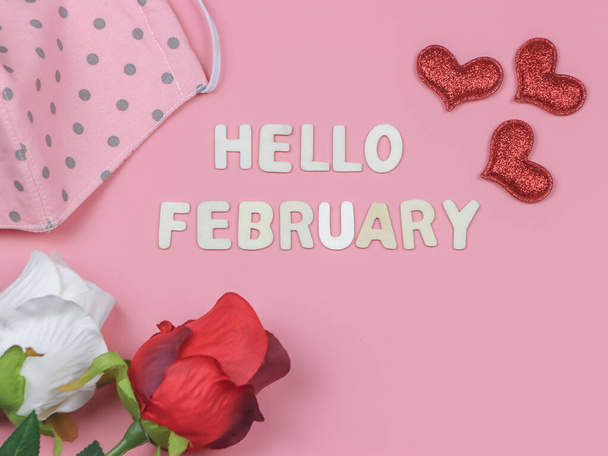 Top view or flat lay of wooden letters HELLO FEBRUARY  with pink polka dots fabric  face mask, red glitter heart, white and red roses on pink background. COVID19, new normal and Valentine's day. - 写真・画像