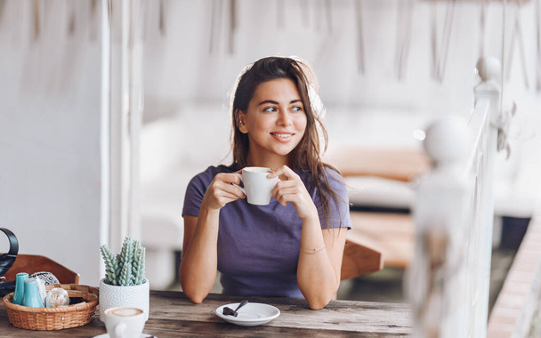 Una chica hermosa y sonriente se sienta en una mesa de madera con una taza de café. - Foto, imagen