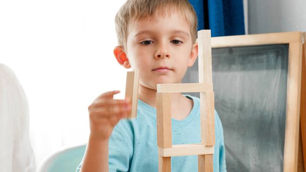 Szczęśliwy uśmiechnięty chłopiec umieszczenie drewnianego bloku na szczycie wysokiej wieży z zabawek drewnianych klocków i cegieł. Koncepcja edukacji dzieci w domu podczas zamknięcia i pozostawania w domu - Zdjęcie, obraz