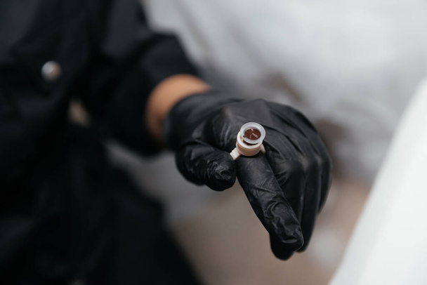 Finger in a black glove is wearing an permanent paint ring with brown pigment. Close-up of pigment container for eyebrow or lip tattoo. Beauty procedure preparation. Selective focus, close-up - Fotó, kép